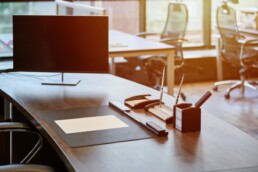Uno spazio di lavoro ufficio vuoto e contemporaneo che presenta una scrivania in legno scuro con un monitor del computer, un elegante tappetino da scrivania nero e forniture per ufficio. Lo sfondo mostra un ufficio open space con luce solare che filtra.