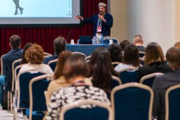 sala conferenze durante un convegno, in primo piano persone sedute rivolte a uno schermo in secondo piano un uomo presenta con un proiettore
